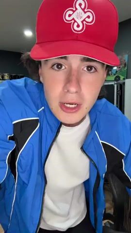 A young person with long, dark hair, wearing a red baseball cap with a white logo, a white t-shirt, and a blue jacket with black and white accents, stands in what appears to be a music studio. The person is looking directly at the camera with an open mouth and animated facial expression. The background shows various musical instruments, including keyboards, drums, and audio equipment, as well as some artwork on the wall. The person begins to dance, moving their arms and legs with energetic gestures.