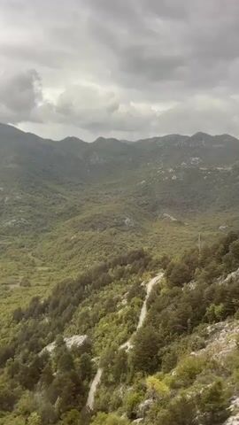 The video depicts a mountainous landscape under an overcast sky. The hills are covered in dense green vegetation, dotted with rocky outcrops. A winding road is visible in the lower portion of the frame. The camera moves, offering a dynamic view of the terrain. A metal railing suggests the perspective is from a moving vehicle, likely a train or car.