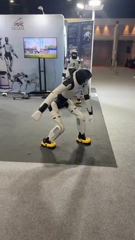 A humanoid robot with a black head and white body is performing a series of dance moves on a dark gray carpeted floor. The robot has yellow feet. In the background, there is a large white booth with a screen displaying images of robots. Another humanoid robot is standing behind the dancing robot. There are also other robots and displays visible in the background. The environment appears to be an indoor exhibition or convention center.