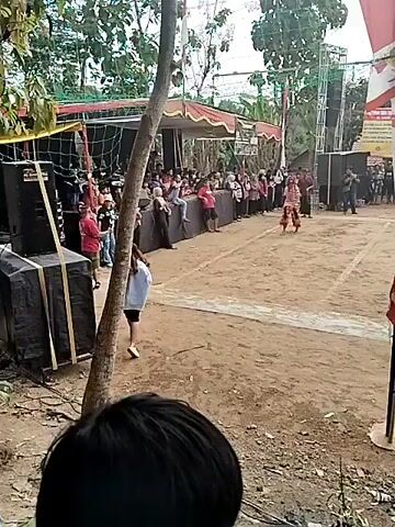 The video shows an outdoor event with a performance taking place on a dirt field. There are people gathered on the sidelines watching. In the center of the field, several individuals are engaged in a performance. One person in the foreground is wearing a striped shirt and black pants. Other performers are wearing traditional costumes, some of which appear to be horse-like. There are trees and buildings in the background, and flags are visible, including what appears to be an Indonesian flag. A volleyball net is set up across the field. The ground is covered in dirt and sand, with some white lines demarcating areas of the performance space. Several tents or awnings are set up along the sides of the field. The lighting suggests it is daytime, possibly overcast. The video captures a dynamic scene with movement and interaction among the performers.