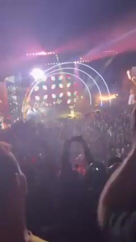 The video captures a live concert from the perspective of a member in the crowd. The stage is brightly lit with colorful lights forming an arch shape. The band is playing, and a large crowd is visible, with many people raising their hands and cheering.  Fog or haze effects enhance the visual atmosphere. The scene is energetic and captures the excitement of the concert experience.