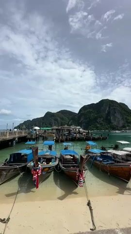 A scenic view of a tropical harbor with numerous traditional longtail boats docked along a sandy beach. The boats are primarily wooden with blue canopies and colorful floral decorations tied to their prows. In the background, lush green, rocky islands rise from the turquoise water. A pier extends into the water, lined with more boats and structures that appear to be part of a small town or market. The sky is partly cloudy with a bright, diffused light, suggesting it is daytime, likely late morning or early afternoon. The water is clear and shallow near the shore, allowing the sandy bottom to be visible. The scene evokes a sense of a popular tourist destination, likely a location for island hopping tours given the presence of many excursion boats. There is no visible text in the image.