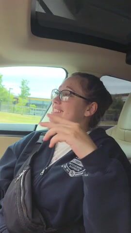 A young woman with glasses and her hair in a bun is sitting in the passenger seat of a car. She is wearing a dark blue hoodie over a white shirt and a black fanny pack. She gestures with her hands and speaks animatedly. The interior of the car is visible, including the headrest of the seat in front of her and the roof of the car with a sunroof. Outside the car window, trees, a fence, and a building can be seen.