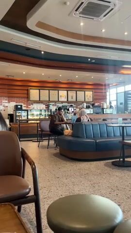 The video shows the interior of a cafe with two people seated at a booth and another person standing behind a counter. The cafe has a modern design with wooden accents, a tiled floor, and a ceiling with exposed ductwork and lights. There are tables and chairs scattered throughout the space, as well as a counter with food and drink displays. The overall atmosphere appears to be relaxed and casual.