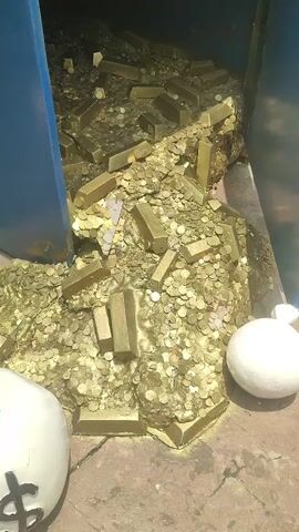 The video shows a themed display with a large pile of gold-colored coins and rectangular bars filling a recessed area. On top of the display are several cartoonish figures: two green characters with cowboy hats and a pink piggy bank, all standing on a mound of the same gold-colored coins. The display is painted with light blue accents and has a playful, child-friendly design. The location appears to be outdoors, possibly in an amusement park or a similar entertainment venue. The camera pans up to reveal the full display after initially focusing on the gold pile.