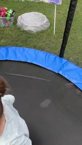 Two young children are playing on a trampoline. The child on the left is wearing a yellow t-shirt and green pants with a dinosaur pattern. The child on the right is wearing a light blue shirt and has their hair pulled up with colorful hair ties. They are both on their hands and knees, and the child on the left appears to be reaching out. The trampoline has a black jumping mat and a blue safety pad around the edge. The trampoline is outdoors, with green grass visible in the background, as well as a fence and a building. The sky is visible in some shots, with clouds present.