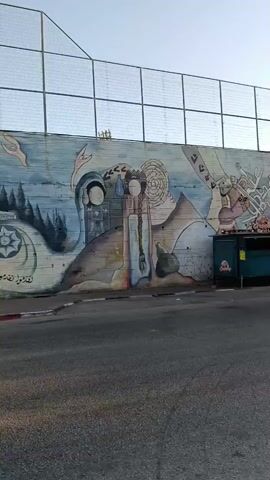 The video pans across a mural painted on a concrete wall. The mural depicts various scenes, including a woman in a headscarf holding wheat, a golden-domed building resembling the Dome of the Rock, and stylized figures. The mural is located next to a road with a curb and a sidewalk. A yellow taxi and a white car are seen driving past on the road. Above the wall, there is a fence with a diamond mesh pattern. The sky is clear and blue. The mural appears to be a significant piece of public art, possibly with cultural or political significance.