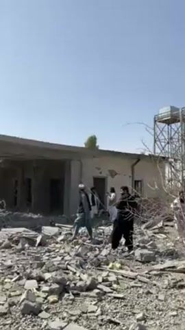 The video shows a group of men inspecting the wreckage of a building. The building appears to have been heavily damaged, with rubble and debris scattered across the ground. The men are dressed in traditional clothing, including turbans and long robes. Some of the men are holding cameras. They are walking around the wreckage, looking at the damage. The sky is clear and blue. It is daytime.