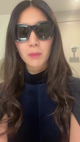 A woman with long, dark, wavy hair is recording a video of herself in what appears to be a bathroom. She is wearing large, black, angular sunglasses and a dark blue, velvet-like top with a high neck. She is looking directly at the camera and talking, gesturing with her hands. The bathroom has a light-colored wall, a partial view of a mirror, and a sink fixture. Music is playing in the background.