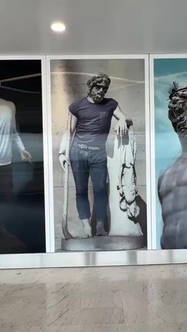 A video shows a digitally altered image of a classical statue of a man. The image is displayed on a wall, likely in a store window, with other images to its left and right. The statue, which appears to be made of marble, depicts a muscular, bearded man holding a staff and leaning on a draped object. The video transitions through several different digitally added clothing items onto the statue. Initially, the statue is shown wearing a t-shirt and jeans. Then, the t-shirt is removed, revealing a bare chest. Next, the jeans are replaced with a thong, and then with boxer briefs. Finally, the t-shirt is put back on, and the boxer briefs are replaced with jeans. The background of the statue's image is a muted, textured wall. To the left of the statue is a partial image of a mannequin or a figure wearing casual clothing. To the right is another image, also appearing to be a digitally altered classical sculpture. The overall environment seems to be an indoor public space, possibly a shopping mall or a retail store, with reflective surfaces and lighting suggesting commercial display.