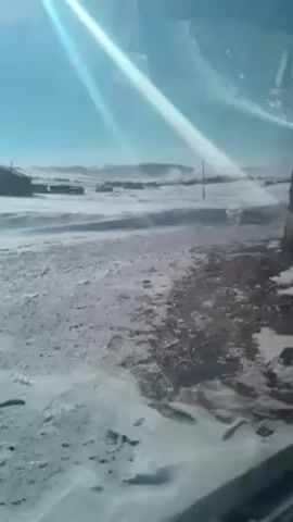 The video depicts a snow-covered landscape under a bright, sunny sky.  A scattering of small buildings is visible in the distance. Snow is being blown across the ground, creating a hazy effect. The foreground shows a mix of snowy and icy patches on what appears to be a road or pathway. Sunlight reflects brightly off the snow and shines through what looks like a vehicle window, causing glare. A building passes by the right of the window.