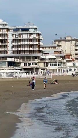 The video captures a beachfront scene with a mix of human activity and architectural elements. The foreground shows a sandy beach with gentle waves lapping at the shore. Several people are scattered across the beach: a woman wearing jeans and carrying a pink bag walks, a shirtless man stands, and a man in a blue shirt holding a dog walks. Some people are lying down, possibly sunbathing. In the background, there are large, multi-story buildings with white balconies and windows. A white fence runs parallel to the buildings, and a bird flies above them. The overall tone of the video is calm and relaxed.