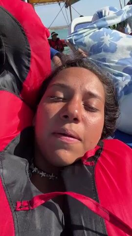 The video shows a woman and a boy lying down on the deck of a boat, both wearing red and black life vests. They are close together, with the woman's face in the foreground and the boy's head resting slightly behind her. The boat appears to be moving, as the ocean and other passengers can be seen in the background. The woman looks directly at the camera. The boy has a camouflage hat. The sun is shining. There appears to be wind.
