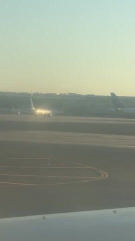 The video shows an airplane on a tarmac with other airplanes in the background. The sun is shining brightly, reflecting off the plane. The sky is clear and pale blue. The tarmac is gray with yellow lines.