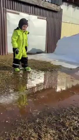 A child in a bright lime green snowsuit and dark gray hat is standing near a large puddle of muddy water on a patch of grass. They are wearing black mittens and lime green rubber boots. Behind the child is a wall made of vertical brown and light yellow planks, with a white door on the left. Next to the wall is a pile of snow. The child stomps in the puddle, causing a splash of muddy water. The camera remains still throughout the short clip.