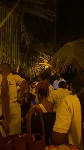 The video shows a group of people gathered at night, possibly at an outdoor event or party. They are mostly wearing white or light-colored clothing. Some are standing, while others are seated at tables. There are palm trees overhead, and lights are strung throughout the area. People are talking, looking around, and generally enjoying themselves. There are bags on the tables. The atmosphere appears lively and relaxed.
