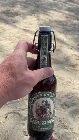 A person's hand is holding a brown glass beer bottle with a swing-top lid. The bottle has a label with the brand name "Kapuziner Weißbier" and an illustration of a monk. The background appears to be a sandy surface, possibly a beach, with some blurred foliage visible. The person's fingers are adjusting the metal clip on the swing-top lid.