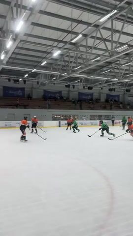 The video shows an indoor ice hockey game. There are two teams, one in orange and black uniforms and the other in green and black uniforms. Several players are visible on the ice, along with hockey sticks and pucks. The game appears to be in progress, with players skating, passing, and potentially shooting the puck. Spectators are visible in the stands in the background. The environment is a standard indoor ice rink with overhead lighting and a metal roof structure.