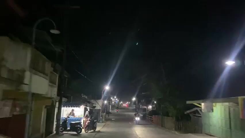 The video shows a view from a vehicle traveling down a street at night. Streetlights illuminate the road, casting long streaks of light. On either side of the street are buildings, some of which appear to be shops or homes. There are a few motorcycles and scooters parked along the side of the road, and one motorcycle is seen moving away from the camera. A food stall with glowing lights is visible on the left side of the street, with a person attending to it. Trees and vegetation are present on the right side of the road, forming a dark backdrop against the night sky. The overall scene depicts a quiet, dimly lit street in what appears to be a residential or mixed-use area at night.