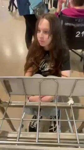 A young woman with long brown hair sits in a metal luggage cart, which is being pushed, in an airport. She is wearing a black t-shirt, light blue pants, and black and white sneakers. The airport concourse has a shiny, light brown floor and is populated with other travelers. In the background, there are signs and chairs.