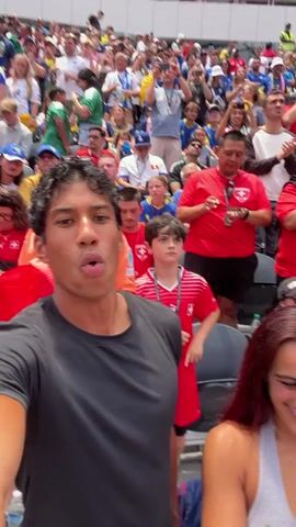 The video begins with a man in a black t-shirt looking directly at the camera with his mouth open in excitement. Behind him, a crowd of people are visible in a stadium. Many are wearing red shirts with white accents, and some are holding Swiss flags. To the man's right, a woman with reddish-brown hair is smiling and looking down. The camera then pans to show more of the stadium, including rows of spectators and signage that reads "FIFA WOR". The man continues to look around the stadium with a mix of excitement and observation. The video concludes with a close-up of the man looking upwards, suggesting he is watching an event unfold in the stadium. The overall atmosphere appears to be one of a sporting event, likely a soccer match, given the "FIFA" signage and the red and white Swiss attire.