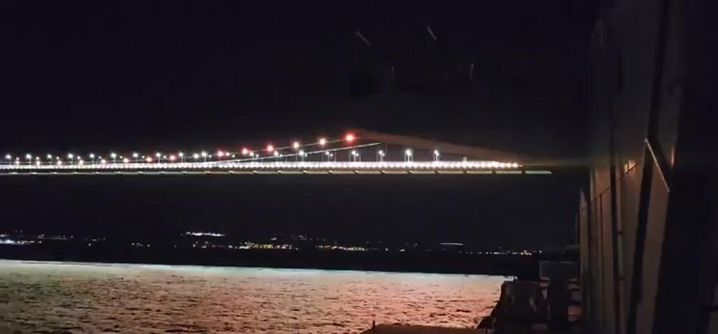 The video shows a suspension bridge at night, illuminated by rows of white and red lights. The bridge spans across a body of water, with lights of a distant city visible on the far shore. The water reflects the bridge's lights, creating a shimmering effect. The camera appears to be on a moving vessel, as evidenced by the slight sway of the image and the way the lights on the bridge appear to streak horizontally. In the foreground, a portion of the vessel is visible on the right side of the frame.