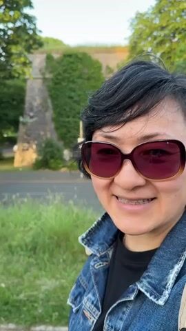 A woman with short, dark hair and wearing large, brown sunglasses, a blue denim jacket, a black t-shirt, and a light beige backpack is seen in a close-up selfie. She is smiling and waving at the camera. The background includes greenery, a road, a stone structure covered in ivy, and trees. The lighting suggests it is daytime.