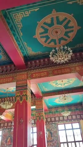 The video pans across the interior of a vibrantly decorated temple. The ceiling is a bright turquoise adorned with ornate gold patterns and large Dharma wheels. Red pillars with intricate gold and green designs support the ceiling. Chandeliers hang from the ceiling, and sconces are mounted on the pillars. Murals depicting Buddhist imagery line the walls. In the center of the frame, a large golden Buddha statue is visible behind an ornate archway, surrounded by many smaller golden Buddha statues. Three men are standing in the foreground. The man on the right is holding a smartphone and appears to be taking a picture or video. The man in the middle wears a straw hat and a black t-shirt, and the man on the left wears a yellow shirt.