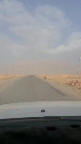 The video shows a view from a vehicle driving on an asphalt road through a desert landscape. The road stretches into the distance, leading towards a hazy horizon. The surrounding terrain is dry and sandy, with sparse vegetation. The sky is partly cloudy, with large white clouds against a blue backdrop. Dust or sand is visible in the air, particularly in the distance, obscuring the view of any mountains or distant features. There are a few utility poles visible along the side of the road. The vehicle appears to be moving forward at a steady pace, as indicated by the slight motion blur and the unchanging perspective of the road ahead.