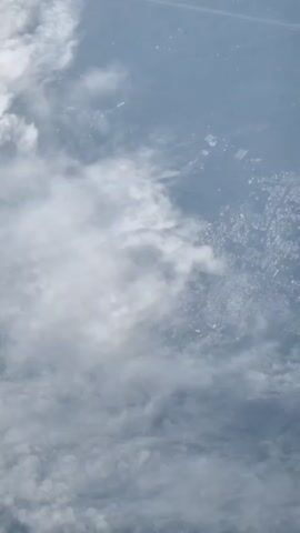 An aerial view of clouds above a city. The clouds are white and fluffy, obscuring much of the city below. The city appears to be a densely populated urban area with many buildings and roads visible. The sky is hazy and overcast, giving the scene a muted, somewhat monochromatic appearance.