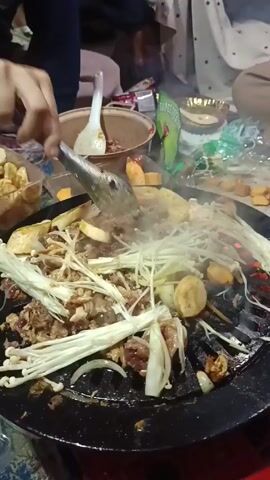 Here's a description of the video:

The video shows a close-up of a person using tongs to stir and cook food on a round, black, sizzling grill. The food includes sliced beef, enoki mushrooms, onions, and other unidentified items. Steam rises from the grill. Various containers of food items surround the grill, including a pot, a plastic container with what appears to be fried food, and a bag of green sauce. Several people sit around the grill, but their faces are not visible. The overall setting seems informal, possibly an outdoor gathering.