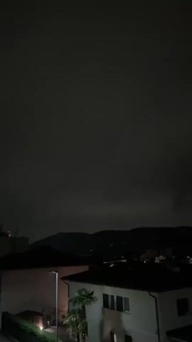 The video shows a nighttime scene with a dark, cloudy sky over a silhouetted landscape of mountains and buildings. A brief flash of lightning illuminates the clouds from 00:01 to 00:03. The foreground shows the rooftops of several buildings, including one with a palm tree in front of it. A streetlamp is visible on the left side of the frame. The overall impression is of a stormy night.