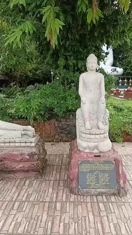 The video shows a panning view of an outdoor Buddhist shrine. There are several white stone statues of Buddha in various poses. In the foreground, a standing Buddha statue is visible on the right, and a reclining Buddha statue is visible on the left. The statues are on pedestals made to look like logs, with inscribed plaques at the base. The ground is paved with stone tiles. In the background, more standing Buddha statues are visible, along with lush greenery, trees, and some structures. A young girl in a light-colored top and blue shorts is briefly seen peeking from behind one of the standing statues. The overall atmosphere appears serene and tranquil.