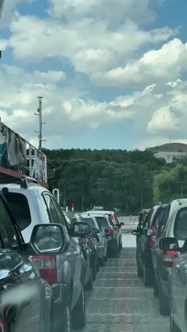 The video shows a view from inside a car waiting on a ferry. The car is one of several lined up on the deck of the ferry. Other cars are visible in front of and behind the car from which the video is being filmed. The ferry appears to be moving or about to move, as indicated by the slight blur and the presence of the water and shoreline in the background. The sky is blue with fluffy white clouds. The ferry has a white cabin with some orange and red accents, and life preservers are visible on the side. The shoreline is lined with trees and green vegetation. The license plate of one of the cars is partially visible and reads "B 4905 BP". The overall scene depicts a typical ferry crossing experience.