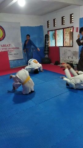 The video captures a martial arts training session in a dojo. The scene is a rectangular room with blue and red mats covering the floor. The walls are painted white and blue, with some areas showing peeling paint. Windows with wooden frames are visible on one side of the room, and Indonesian flags are displayed outside and inside. A punching bag is placed against the wall.

Several individuals, dressed in martial arts uniforms (gi), are engaged in various activities. Some are sparring or practicing techniques, while others appear to be observing or resting. One person in a brown gi is seen on the floor. Another individual in a white gi with a blue belt is on their knees. A person in a white gi with a green belt is standing. The overall atmosphere appears to be one of focused training and practice.

The lighting suggests it is daytime, possibly late afternoon given the shadows. The environment seems to be an indoor training facility. Visible text includes "JAT INDONESIA" on a banner or poster on the wall, and potentially some text on the uniforms, though it's difficult to read clearly. The participants are all young males. The activity is clearly martial arts practice, likely a form of grappling or striking, given the poses and interactions.