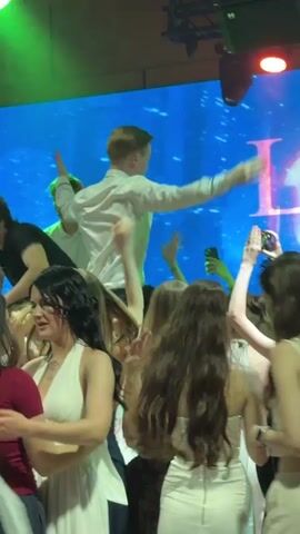 This media file captures a lively scene at what appears to be a celebration or party, possibly a school event like a prom or graduation, given the attire of some individuals. The setting is indoors, with a large LED screen in the background displaying a light blue abstract design with a large pink "L" visible. Stage lighting illuminates the scene with vibrant green and some orange hues.

The primary subjects are a group of young people, a mix of males and females, clustered together. In the center of the frame, a young man in a white dress shirt is being carried on the shoulders of another person, his arms outstretched as if addressing the crowd or reveling in the moment. He appears to be looking to his right. Other young women are visible in the foreground and midground, some in white dresses or outfits, and others in darker clothing. One young woman in a white halter top is looking directly at the camera, while another with dark hair and a red top is looking down. Many individuals have their phones out, suggesting they are capturing the event.

The overall mood is energetic and festive, with people actively engaged and enjoying themselves. The lighting and the presence of a crowd suggest an evening or nighttime event. There are no explicit weather cues as it is an indoor setting. The location context provided is Khotianivka, Ukraine, which could be a clue to the specific event or venue if it's a known local spot. No other significant objects are immediately discernible beyond the people, clothing, and the background screen and lighting equipment.