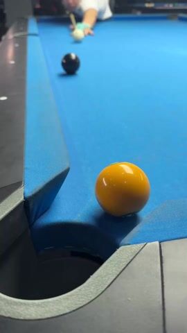 The video shows a person playing pool. The camera is positioned at the corner pocket of a blue pool table, focusing on a yellow ball near the edge. In the background, another ball, black, sits farther up the table. The person takes a shot, hitting the yellow ball away from the corner and pocketing the black ball.