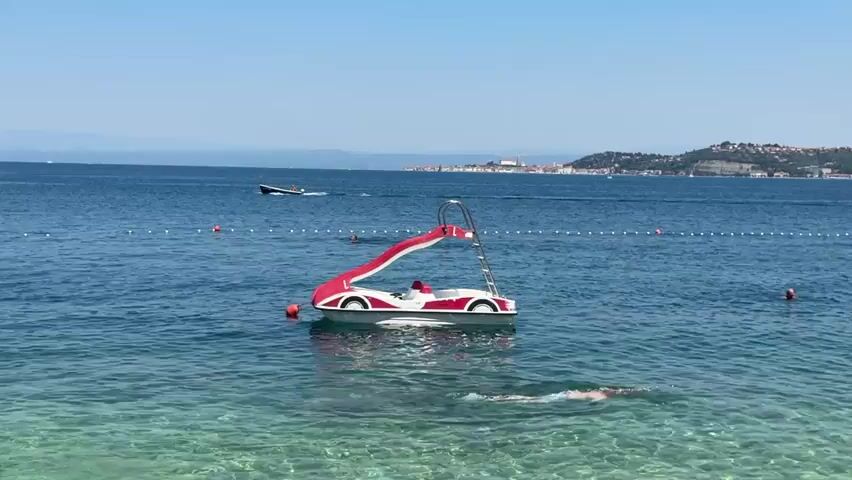 The video depicts a clear, sunny day at a beach with calm, clear turquoise water. In the foreground, a person is swimming towards the right of the frame. Further out in the water, a red and white pedal boat shaped like a car, complete with a slide, is docked by a buoy. Several other people are visible in the water, either swimming or relaxing.

In the background, a speed boat is moving across the water, leaving a wake. A rope with buoys stretches across the sea, marking a boundary. The distant shore is lined with buildings, suggesting a town or resort area, with hills rising behind it. The overall atmosphere is serene and leisurely, indicative of a summer vacation. The clear sky and bright sunlight suggest it is midday. The location, Crveni Vrh, Croatia, is known for its coastal beauty. There is no visible text in the video.