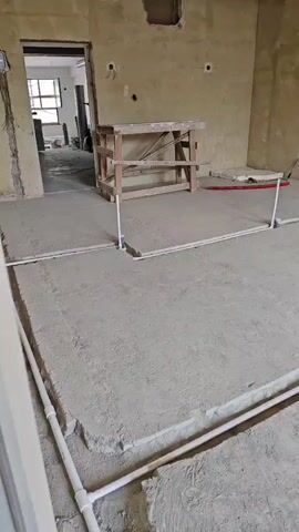 A video captures a construction site. The scene focuses on the floor, which is a bare concrete slab with trenches dug into it. White plastic pipes are laid within these trenches, forming a grid-like pattern across the floor. Some pipes are bent at right angles, indicating a planned layout. In the background, there are unfinished walls with exposed concrete and plaster. A wooden workbench or scaffolding stands in the middle ground, and an open doorway reveals a glimpse of another room with windows and more construction materials. The lighting is natural, suggesting daylight. The overall impression is that of ongoing plumbing or underfloor heating installation. There are no people visible in the video.