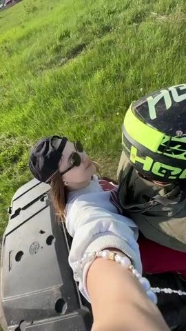 A first-person view video shows a woman with long brown hair, wearing a black cap, sunglasses, a white sweater, red pants, and a pearl bracelet, riding as a passenger on a black ATV. The driver, wearing a green jacket and a neon yellow and black helmet, is operating the ATV on a dirt road.  The road is surrounded by green grass, trees, and a cloudy sky. A white SUV is seen driving on the same road ahead of them. The woman briefly turns toward the camera, smiles, and then looks forward.