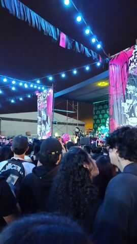 Here is a concise description of the video:

The video shows a crowd of people at an outdoor concert at night. The band, ENO MERCÍANA, is performing on a stage lit with blue and pink lights. Many people in the crowd are recording the performance with their phones. There are strings of lights overhead and a building visible in the background.