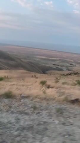 This is a video of a landscape, likely taken from a moving vehicle. The scene is dominated by rolling hills covered in dry, golden grass and scattered bushes. In the distance, there is a wide, flat expanse of land that appears to be a valley or plains. The sky above is a pale blue with some wispy clouds, suggesting it might be early morning or late afternoon. The camera is angled slightly downwards, giving a sense of looking over the landscape. There are no people or identifiable objects visible in the scene. The overall impression is one of a dry, open, and somewhat desolate environment. The video's motion blur suggests a journey through this landscape.