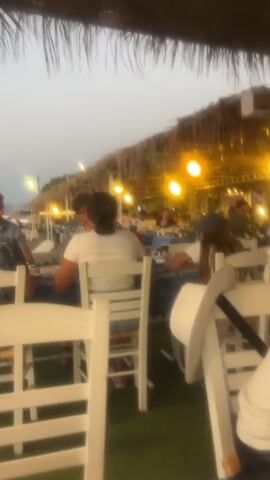 The video shows a blurry outdoor scene at dusk or dawn. In the foreground, white chairs are visible, suggesting a seating area for a restaurant or beachside establishment. Beyond the chairs, people are seated at tables, some with blue tablecloths. The background reveals a beach with lounge chairs and umbrellas, leading to the ocean. The sky is a pale, dusky color, and string lights are illuminated, casting a warm glow. The overall impression is of a relaxed, possibly dining, atmosphere by the sea.