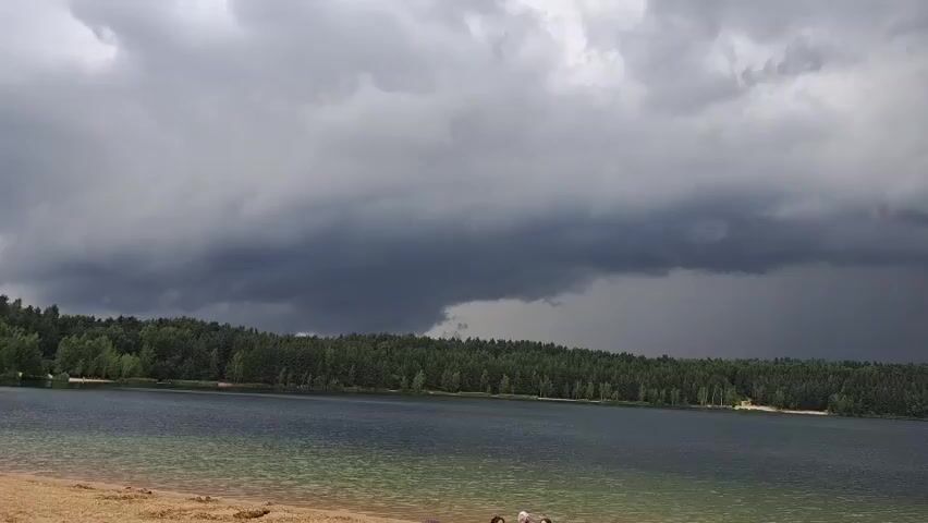 The video shows a wide shot of a lake with a dense forest lining its far shore. The sky is dramatic and overcast with heavy, dark storm clouds. A visible rain shaft is present in the distance, indicating an ongoing or approaching storm. The foreground features a sandy beach area with some indistinct figures visible, and the lake's water is a dark blue-green, appearing calm despite the stormy weather. The overall mood is somber and possibly a bit ominous due to the weather conditions. The location appears to be a natural recreational area, possibly a beach or park, given the presence of the sand and the treeline. There are some small structures visible along the treeline in the distance, which could be cottages or beach facilities. The time of day is unclear but the dark clouds suggest it is not midday with bright sunshine. There is no visible text in the video.