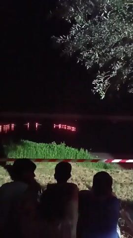 The video shows three people from behind, seated on a grassy area at night, facing a body of water. The water reflects rows of red lights. There is green vegetation along the near bank of the water, and a red and white striped tape is visible in the foreground, suggesting a barrier. A tree with leaves is visible in the upper right corner of the frame. The overall scene is dark, with the primary light source being the reflected red lights on the water.