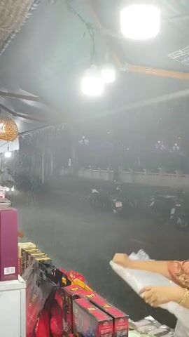 The video shows a street scene at night during a heavy downpour. Rain is falling intensely, creating a misty effect and obscuring some details. There are several motorcycles parked on the side of the road, and at least two individuals are seen riding motorcycles. One rider, wearing a light-colored jacket, is on a motorcycle approaching from the left, their headlight visible. Another motorcycle rider, wearing a yellow rain jacket, is seen further down the road. In the foreground, under a covered area with bright lights, an older woman with gray hair, wearing a patterned blouse, is interacting with some items, possibly products in boxes, on a counter. The environment appears to be a roadside establishment or market stall, with woven materials visible on the ceiling and multiple hanging lights illuminating the area. Decorative purple lights are visible in the background, adding a touch of color to the dark, rainy night.
