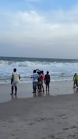 The video shows a group of people on a beach. One person stands away from the group, facing the ocean, with their back to the camera. The group of five are standing closer together at the water's edge, some are interacting, with one putting his hand on another's head. The ocean is visible in the background with waves breaking on the shore. The sky is overcast with some blue patches. There are a few other individuals further up the beach, towards the right side of the frame.