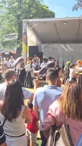 A group of people are on a stage, possibly performing a traditional dance or reenactment. They are dressed in historical clothing. In the foreground, there is a table with bread on it. A crowd of people are watching the performance from the audience. The scene appears to be outdoors, with trees and a stage structure visible. The overall atmosphere suggests a cultural event or festival.
