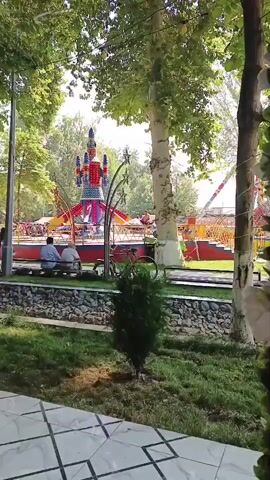 The video shows an amusement park with a rocket-themed ride in the center. Several people are visible sitting on a bench in the foreground, and others are walking around the park. The ride is colorful and appears to be in operation, with people on it. Trees and grass surround the park, creating a natural and park-like setting. The overall impression is that of a pleasant and lively park atmosphere.