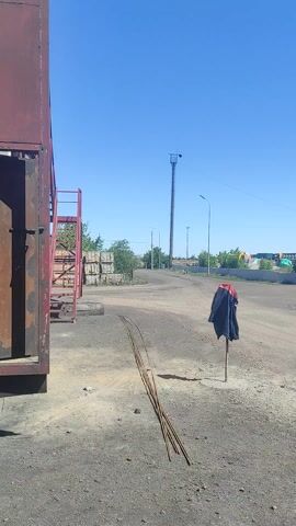 This is a video file recorded on a bright, sunny day with a clear blue sky. The camera appears to be handheld and is tilted vertically, causing the scenes to be sideways. The setting seems to be an industrial or storage area, possibly a refinery or depot, given the presence of large cylindrical tanks, metal structures, a crane, and what looks like construction materials such as rebar and stacked bricks. The ground is covered in gravel and dirt.

In the first segment, a scarecrow made from a dark jacket and a red shirt tied to a pole is visible in an open, dusty area. Further down the road, there are power lines and tall metal towers. In the second segment, a line of large, round, concrete structures are prominent, possibly storage tanks. In the third segment, a dark, large industrial structure with scaffolding is seen behind a fence. The final segment shows more industrial buildings and trees, with the sun glinting brightly in the sky, suggesting it is midday. There are no people visible in the footage. The overall impression is one of an operational or disused industrial site under clear weather conditions.